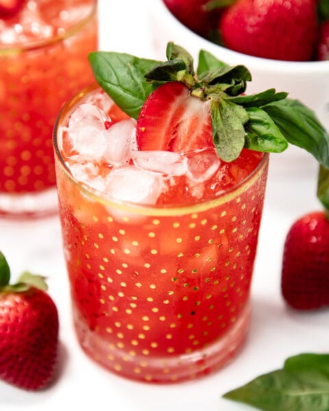 A glass of red strawberry drink with ice, garnished with a halved strawberry and basil leaves, sits beside fresh strawberries and basil on a white surface.