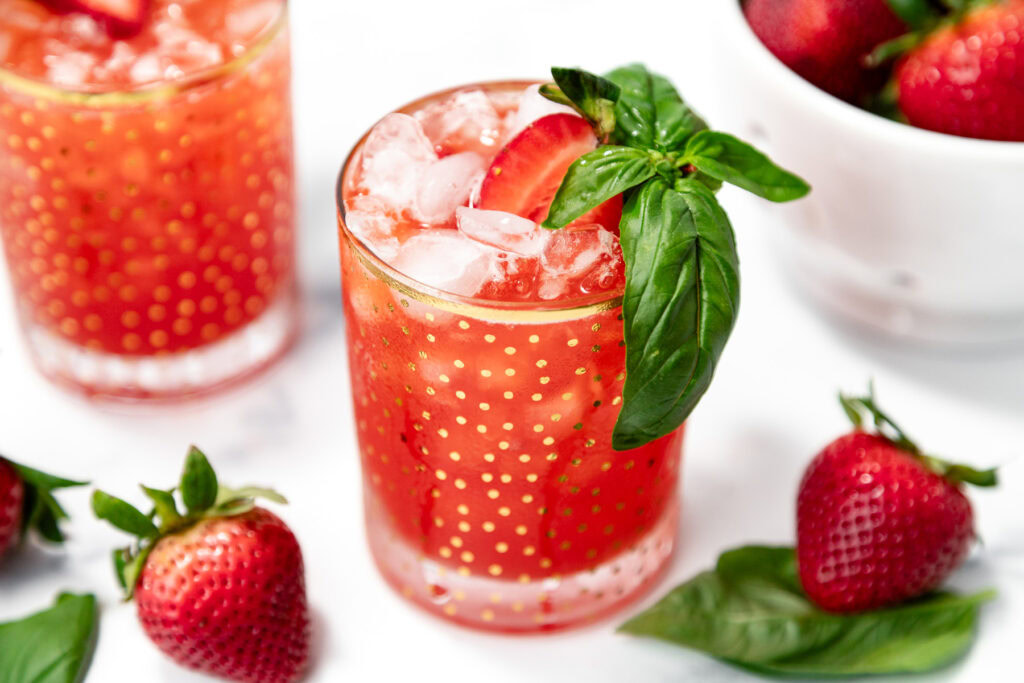 A glass of iced strawberry drink garnished with a fresh basil sprig and a strawberry slice, surrounded by strawberries and basil leaves.