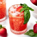 A glass of red iced drink garnished with a basil sprig and plum slice, with strawberries and basil leaves nearby.