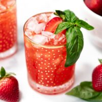 A glass of red iced drink garnished with a basil sprig and plum slice, with strawberries and basil leaves nearby.