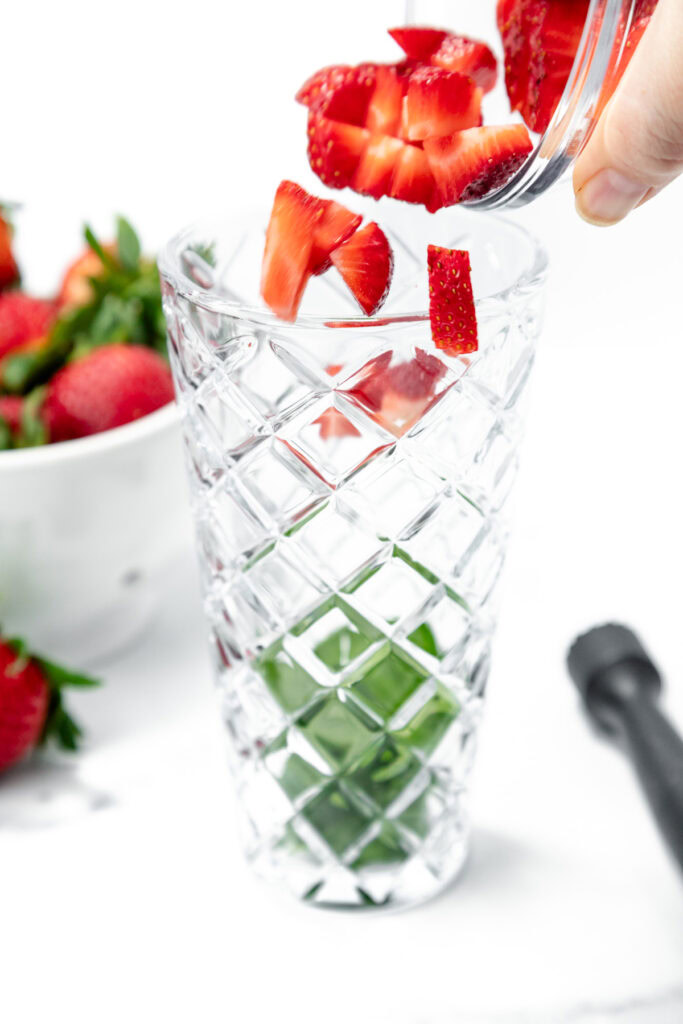 Hand adding chopped strawberries into a patterned glass with mint leaves at the bottom; a bowl of whole strawberries is in the background.