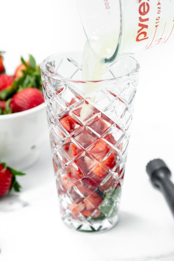 A glass filled with sliced strawberries is being poured with liquid from a measuring cup, with a bowl of whole strawberries in the background.