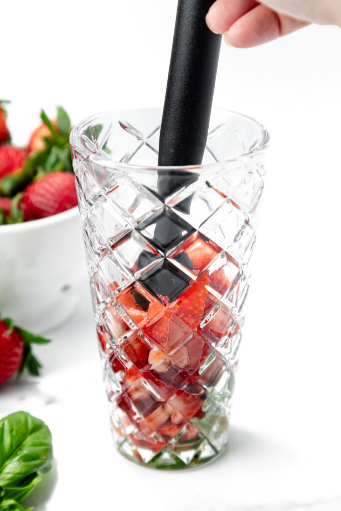 A hand uses a muddler to crush strawberries in a patterned glass; fresh strawberries and basil leaves are visible nearby.