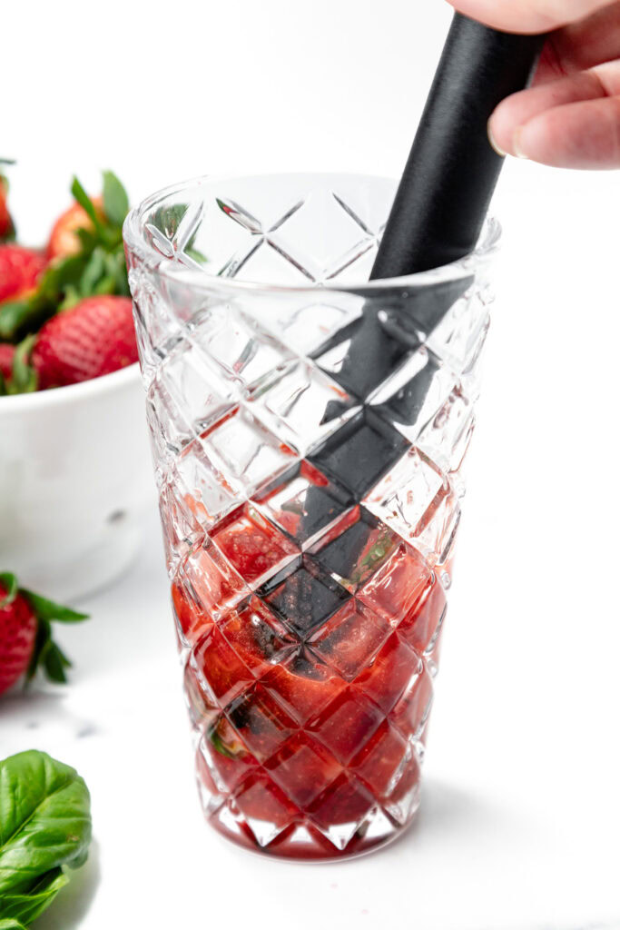 A hand uses a muddler to crush strawberries and basil in a patterned glass; fresh strawberries and basil are nearby on a white surface.