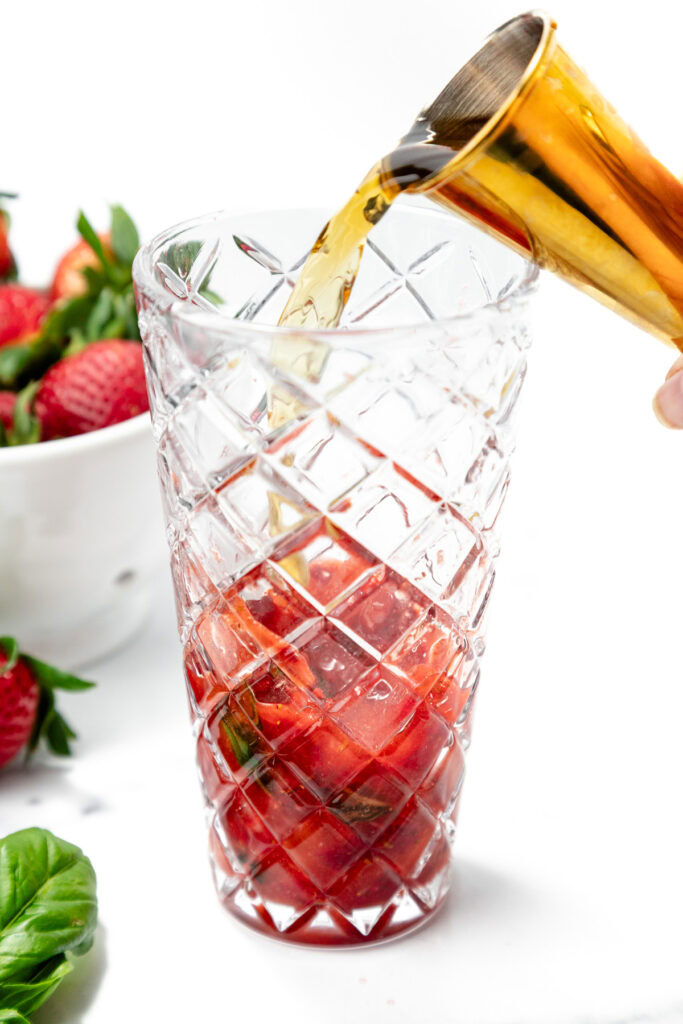 A gold jigger pours liquid into a glass containing sliced strawberries and herbs, with whole strawberries and basil nearby.