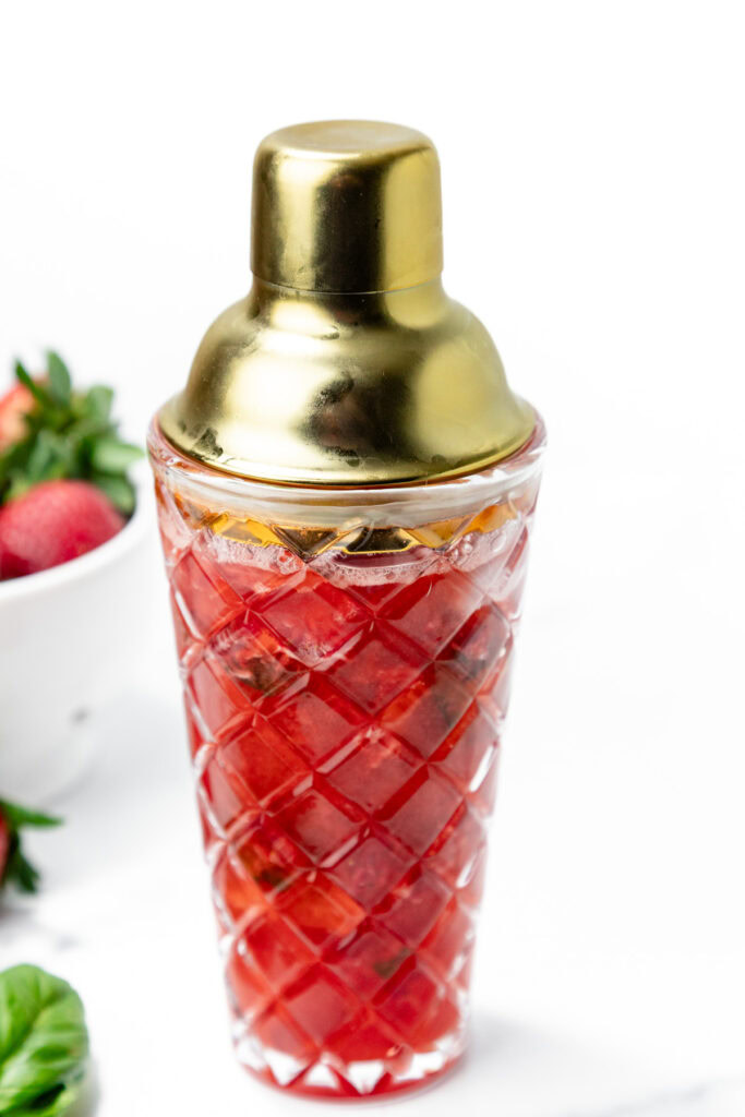 A glass cocktail shaker with a gold lid filled with a red, ice-filled beverage, with strawberries and basil visible in the background.