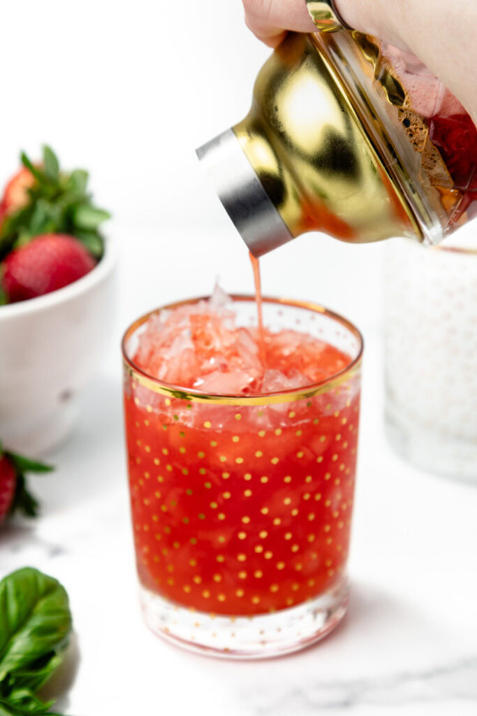 A hand pours a red, iced drink from a gold cocktail shaker into a clear glass with gold polka dots; strawberries and basil are nearby.