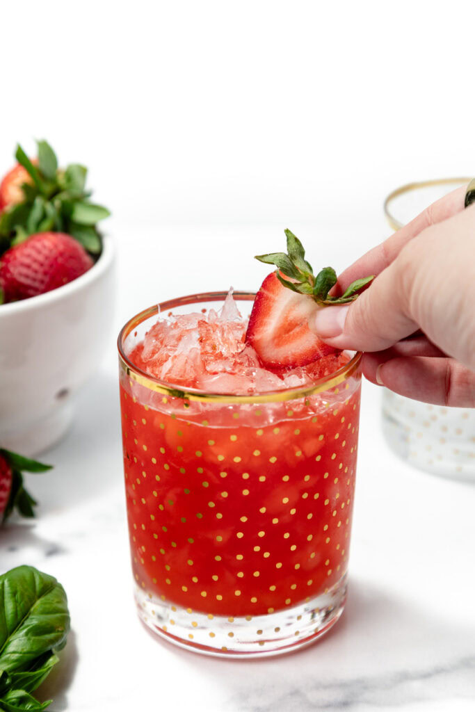 A hand places a sliced strawberry garnish on a glass of iced strawberry drink, with a bowl of strawberries in the background.