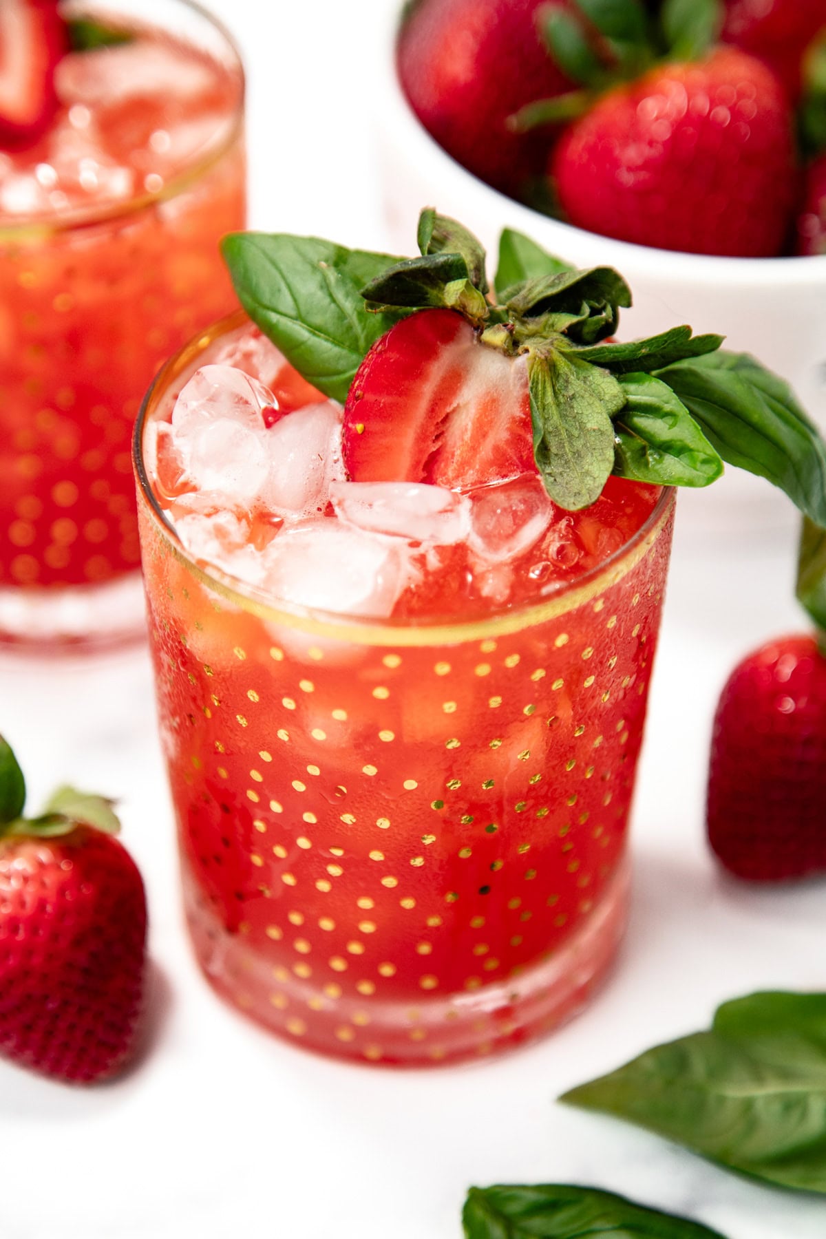 A glass of red strawberry drink with ice, garnished with a halved strawberry and basil leaves, sits beside fresh strawberries and basil on a white surface.