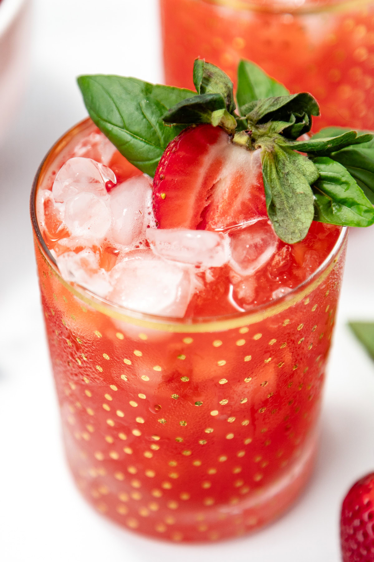 A glass filled with a red, iced beverage garnished with a fresh strawberry and basil leaves, served in a clear glass with gold polka dots.