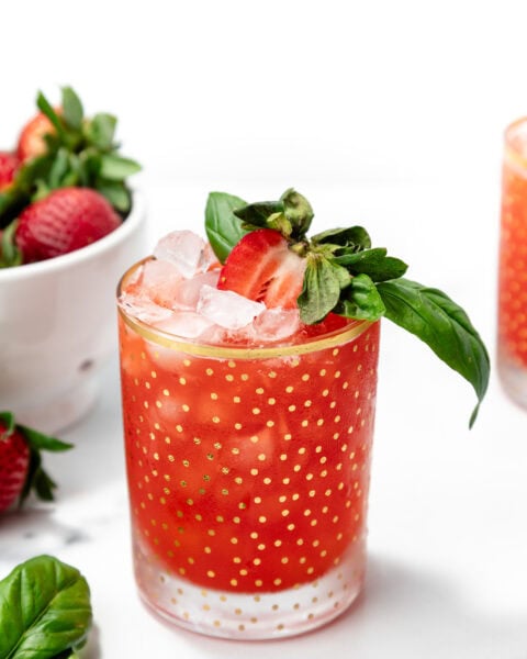A glass of red strawberry cocktail with ice, garnished with a halved strawberry and basil, sits on a white surface next to a bowl of strawberries and basil leaves.