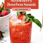 A glass of strawberry bourbon smash with ice, garnished with a fresh strawberry and basil, sits on a white surface beside a bowl of strawberries.
