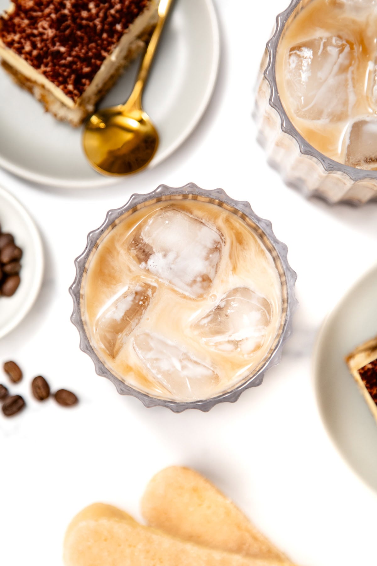 A glass of iced coffee with milk sits on a white surface, surrounded by slices of cake, coffee beans, a gold spoon, and ladyfinger biscuits.