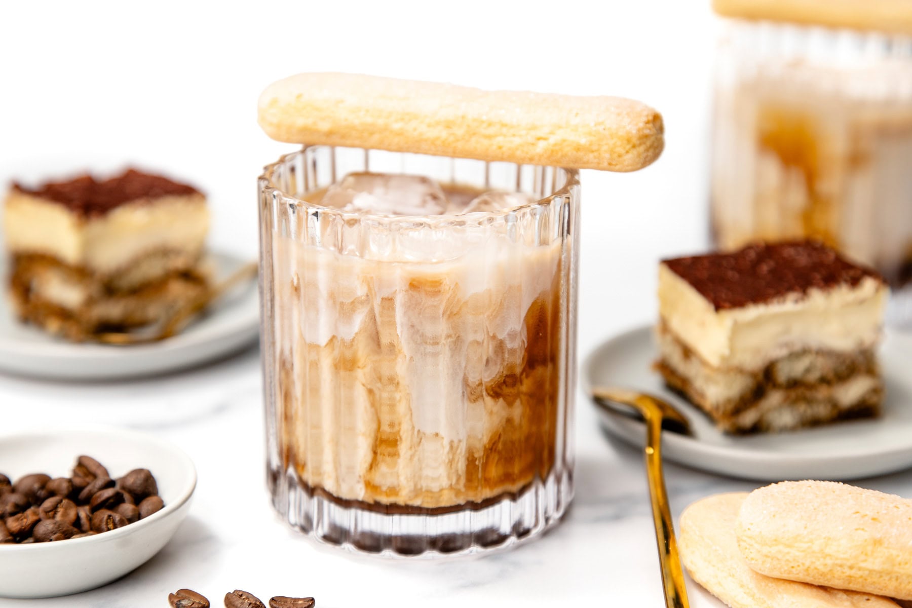 A glass of iced coffee with milk, topped with a ladyfinger biscuit, is surrounded by tiramisu slices, coffee beans, and additional ladyfingers on a white surface.