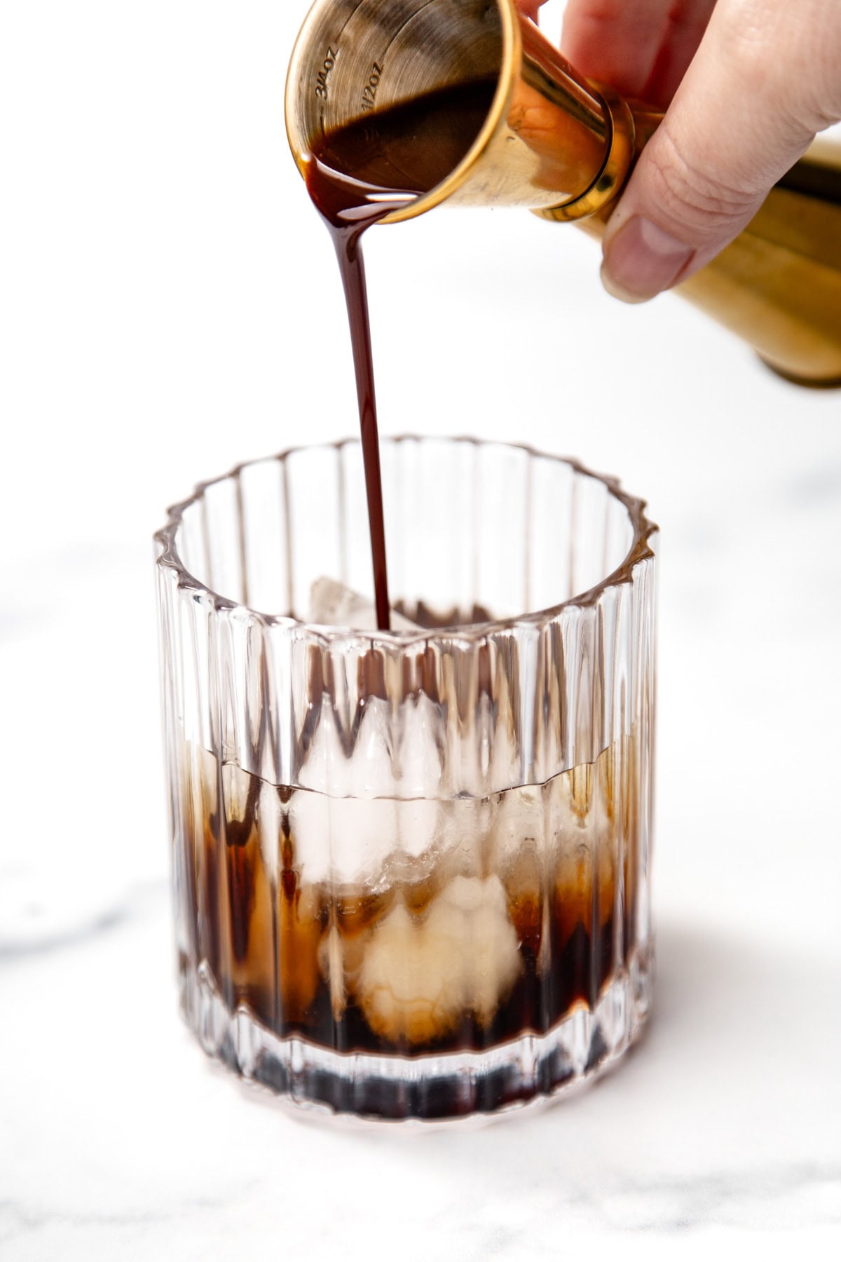A hand pours dark liquid from a jigger into a glass filled with ice on a white surface.