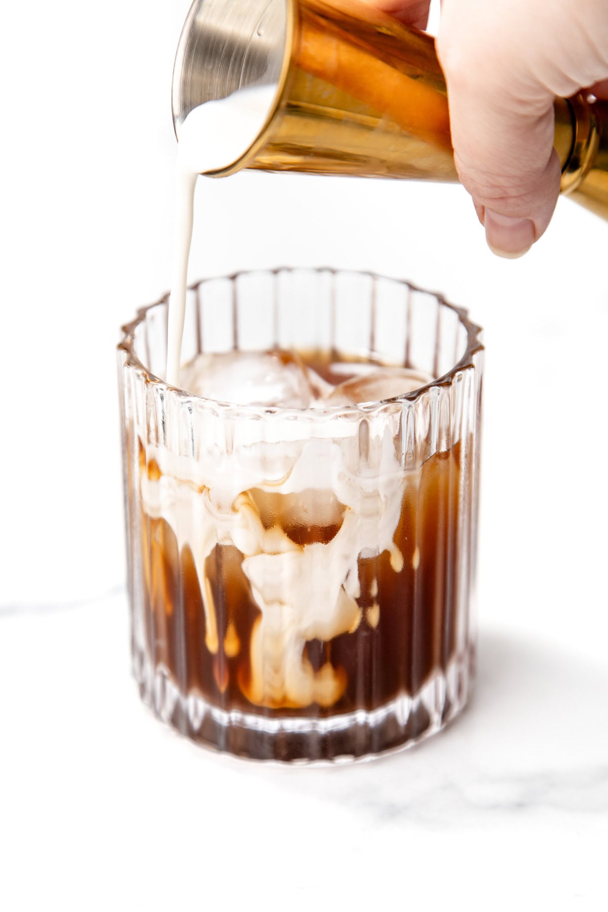 Cream is being poured from a gold jigger into a glass of iced coffee, with the cream swirling through the dark liquid.