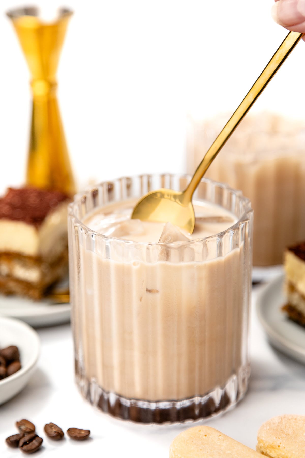 A hand stirs a creamy iced coffee drink in a glass with a gold spoon, surrounded by coffee beans, ladyfingers, and pieces of tiramisu dessert.
