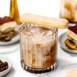 A glass of iced coffee with milk, topped with a ladyfinger biscuit. Plates with tiramisu and a jigger are in the background on a white surface.