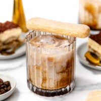 A glass of iced coffee with milk, topped with a ladyfinger biscuit. Plates with tiramisu and a jigger are in the background on a white surface.