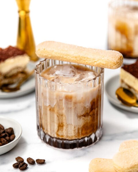 A glass of iced coffee with milk, topped with a ladyfinger biscuit. Plates with tiramisu and a jigger are in the background on a white surface.