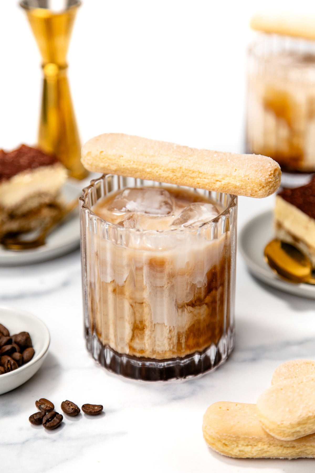 A glass of iced coffee with milk, topped with a ladyfinger biscuit. Plates with tiramisu and a jigger are in the background on a white surface.