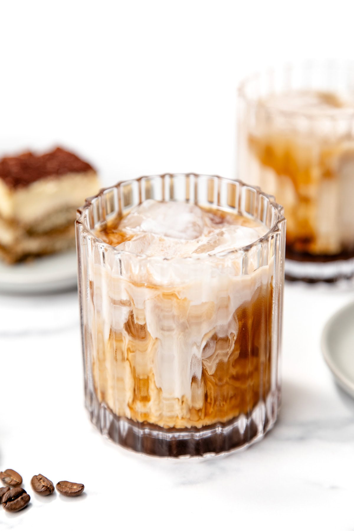 A glass of iced coffee with milk swirling on top, next to coffee beans and a dessert in the background on a white surface.