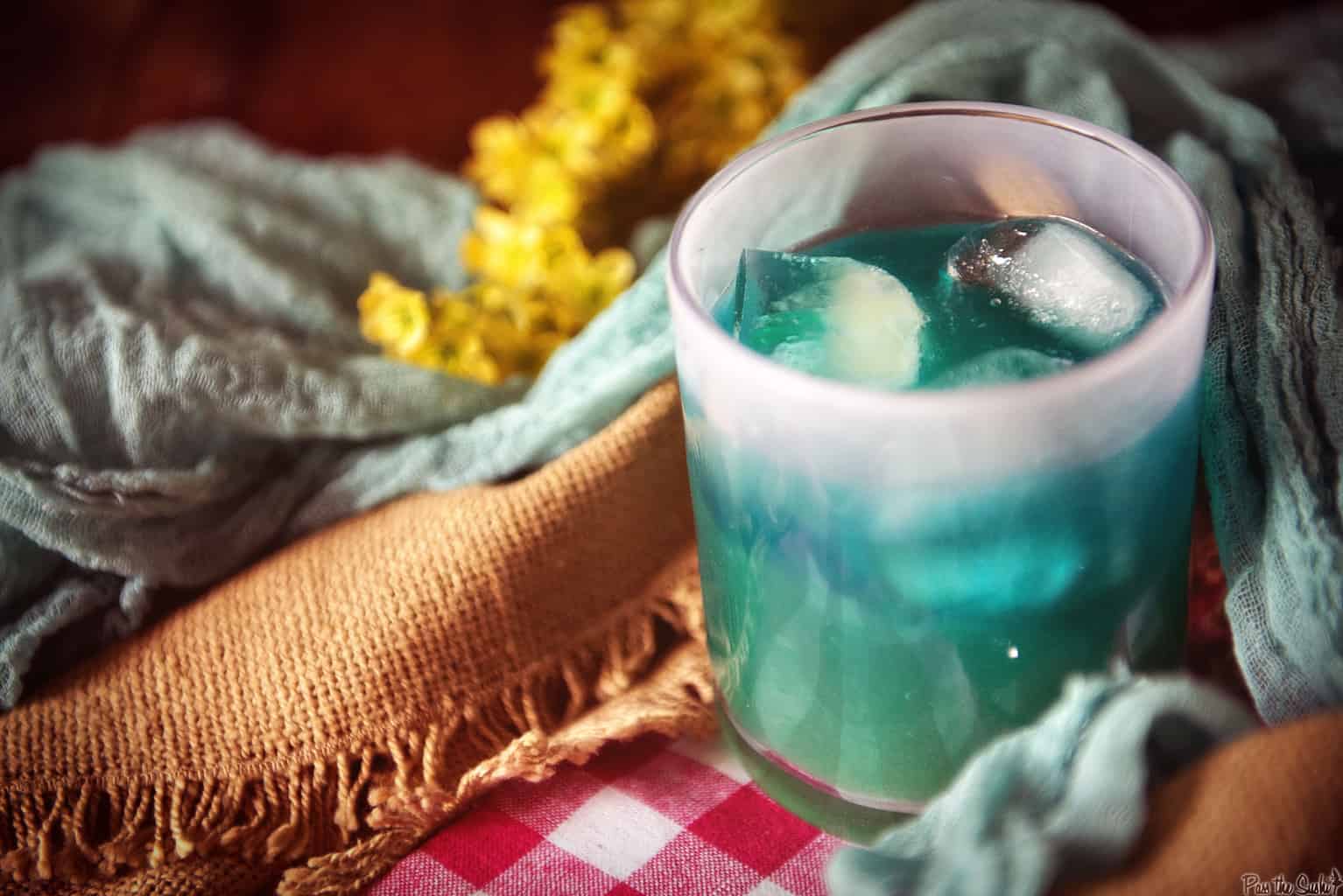 A glass of blue cocktail with ice, set on a colorful cloth with a yellow flower in the background.