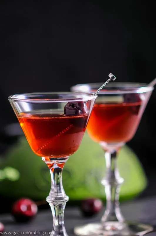 Two cocktails in martini glasses, each garnished with a dark cherry on a cocktail pick.