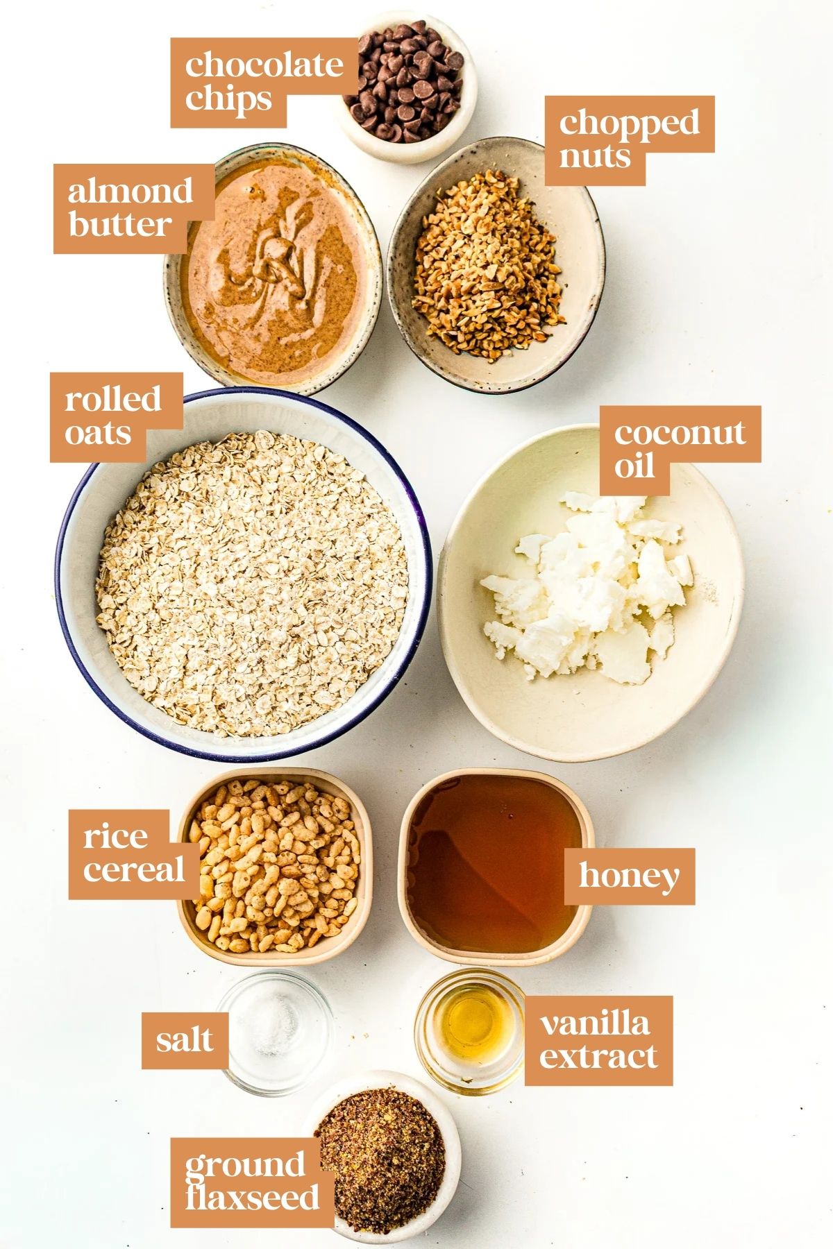 Bowls with labeled ingredients: chocolate chips, chopped nuts, almond butter, oats, coconut oil, rice cereal, honey, salt, vanilla, flaxseed.
