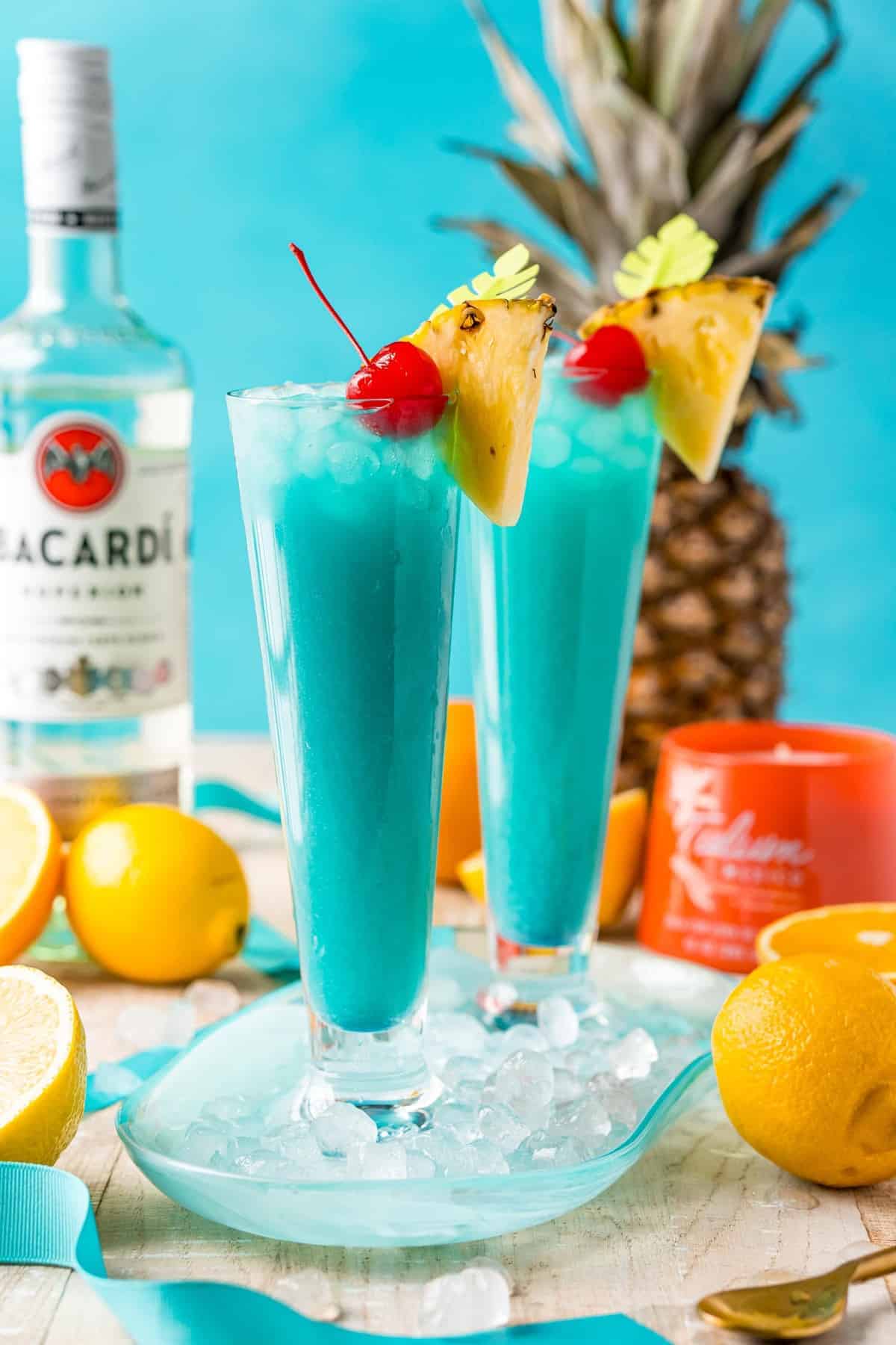 Two tall glasses of blue tropical cocktails garnished with pineapple and cherry, surrounded by fruit and Bacardi bottle.