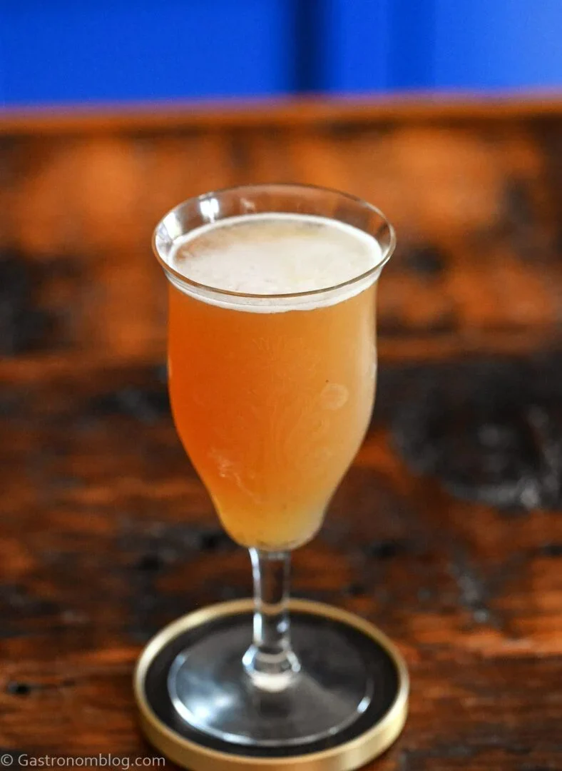 A frothy orange cocktail in a stemmed glass sits on a round coaster atop a wooden table, making it a perfect addition to your Father's Day cocktails lineup.