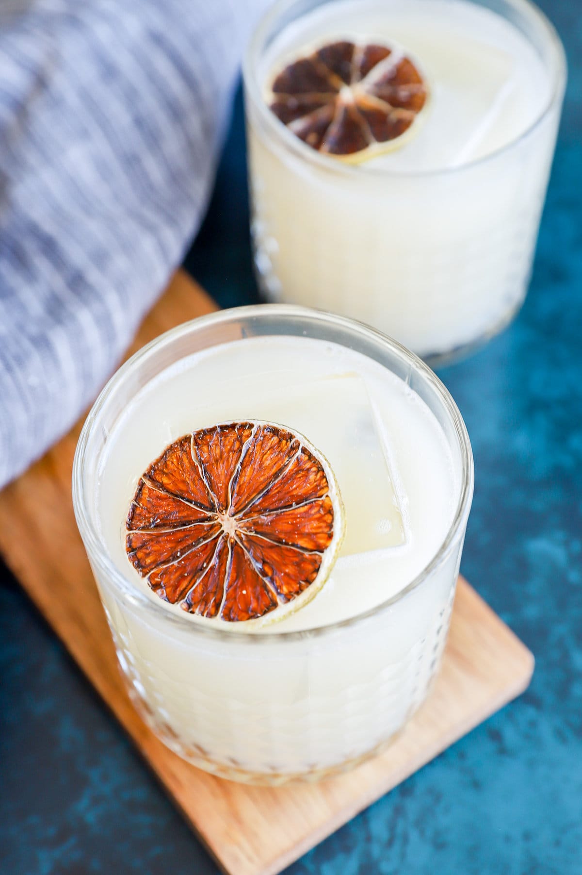 Two glasses of a white drink with large ice cubes, garnished with a dried citrus slice, on a wooden board.