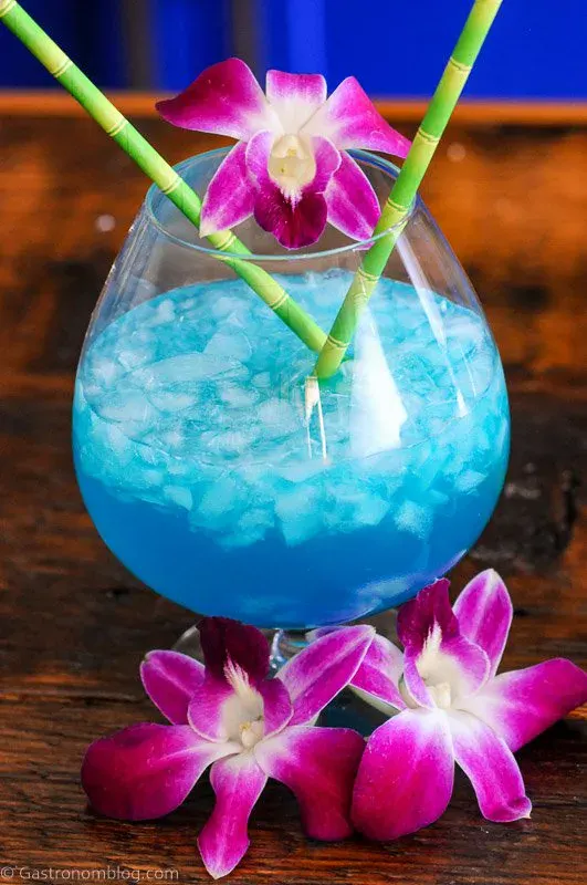 A stunning blue cocktail with crushed ice, part of the latest blue cocktails trend, garnished with orchids and bamboo straws, sits elegantly on a wooden surface.