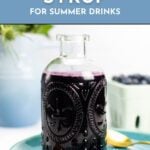A glass bottle of blueberry syrup on a blue plate with fresh blueberries in the background.