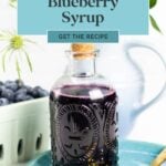 A glass bottle of blueberry syrup on a blue plate, with fresh blueberries and a plant in the background.