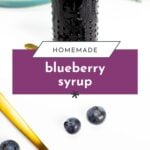 A bottle of blueberry syrup, a gold spoon with syrup, and fresh blueberries on a white surface.