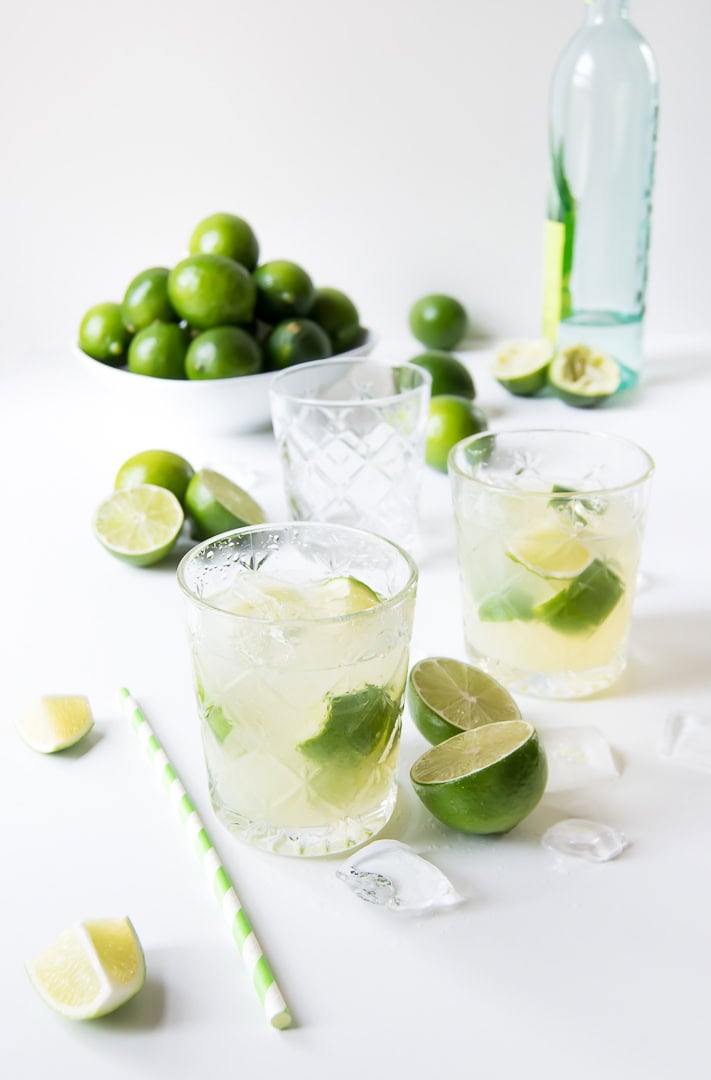 Two glasses of lime cocktails with ice, fresh limes, a bottle, and straws on a white surface.