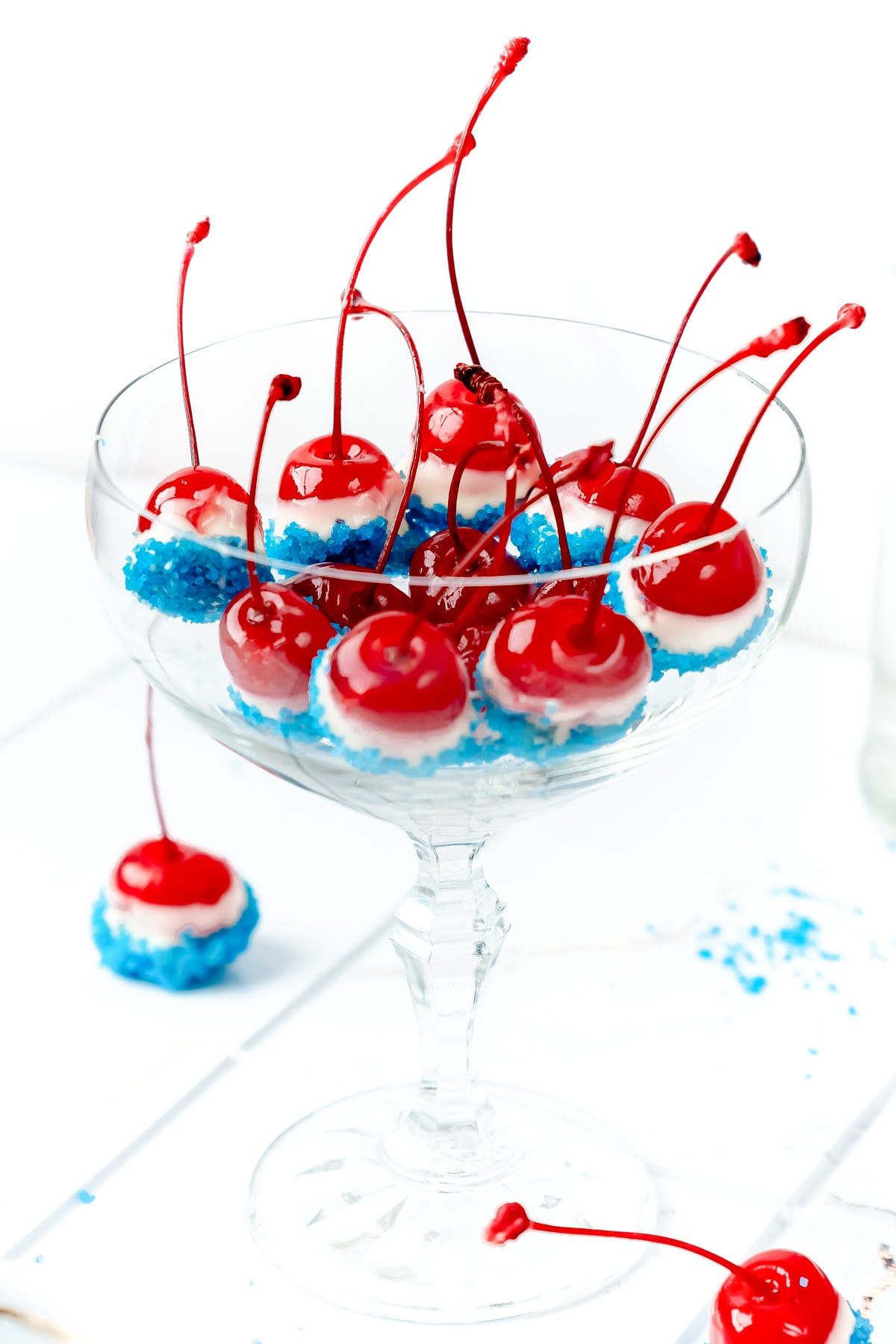 A glass dish filled with cherries dipped in blue sugar, with long stems, on a white background.