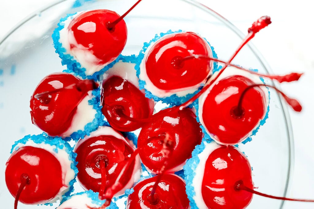 Bright red cherries dipped in white chocolate and blue sprinkles, arranged closely on a white background.