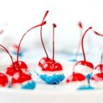 Red cherries dipped in white chocolate and blue sprinkles, arranged on a white plate for a festive treat.