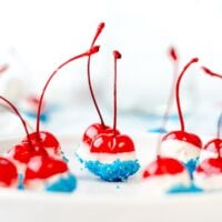 Red cherries dipped in white chocolate and blue sprinkles, arranged on a white plate for a festive treat.