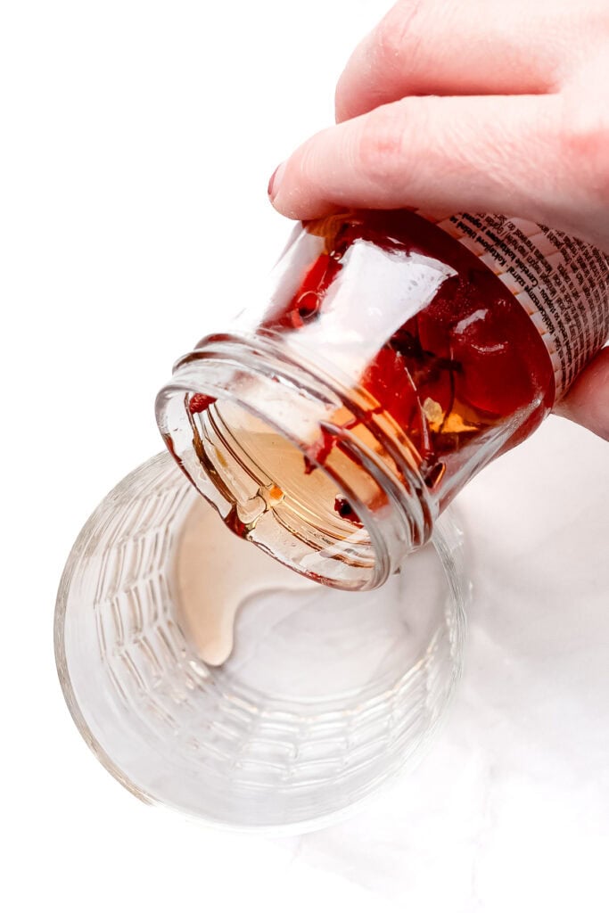A hand pours oil from a glass jar with chili flakes into an empty glass on a white surface.