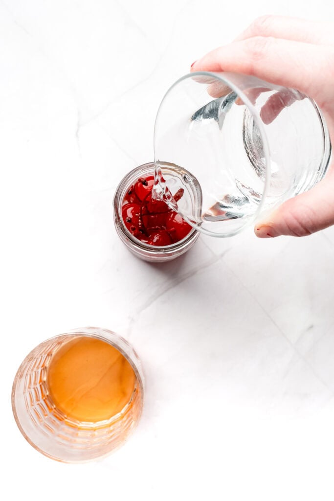 A hand pours clear liquid into a glass with cherries, next to a glass of amber-colored liquid on white surface.