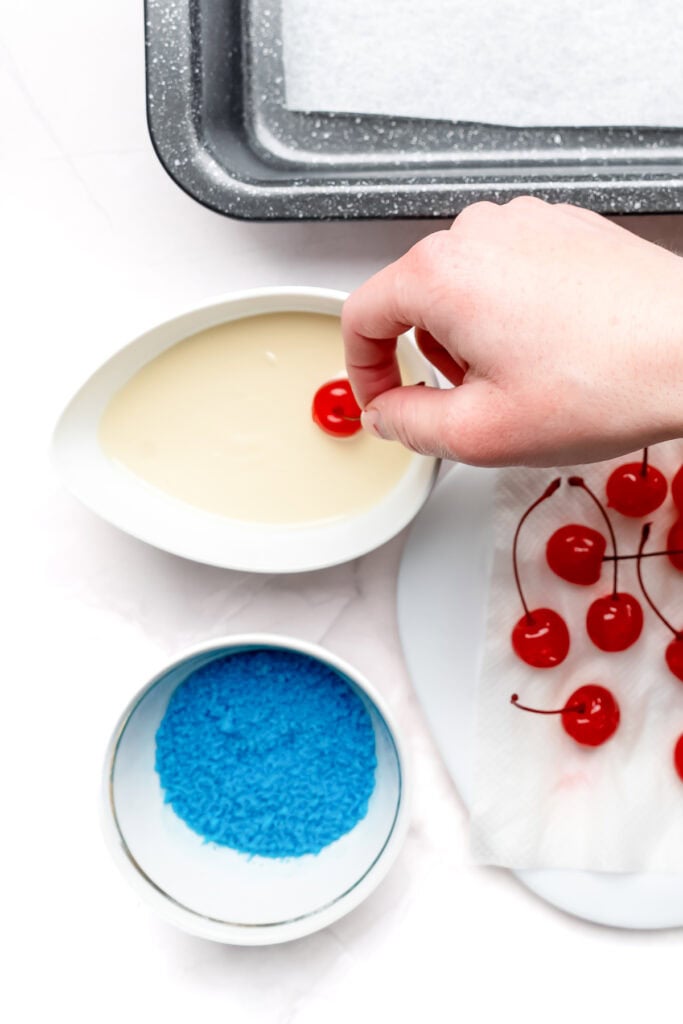 A hand dips a maraschino cherry in white chocolate, with blue sprinkles and more cherries nearby.