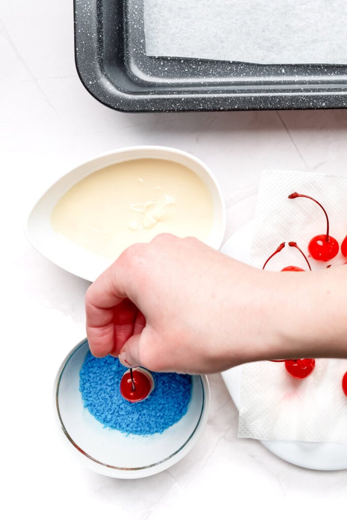 A hand dips a cherry coated in white chocolate into blue sprinkles, with baking supplies nearby.