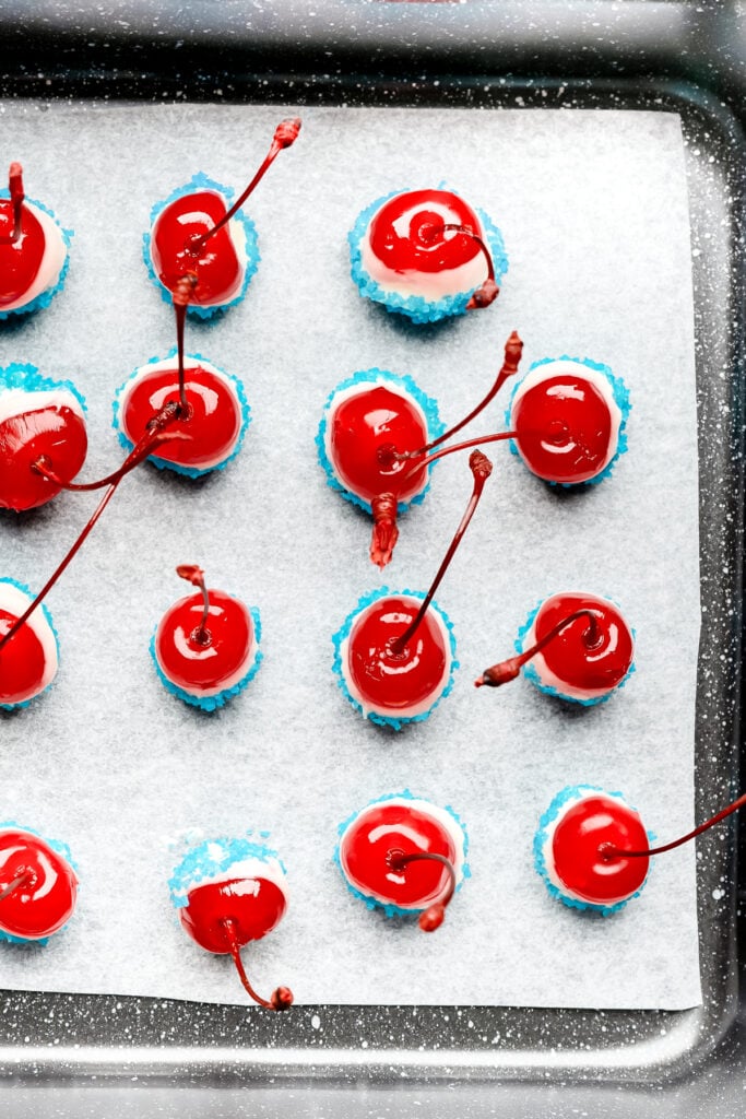 Red cherries with stems on white and blue coated candies, arranged on parchment paper on a baking sheet.