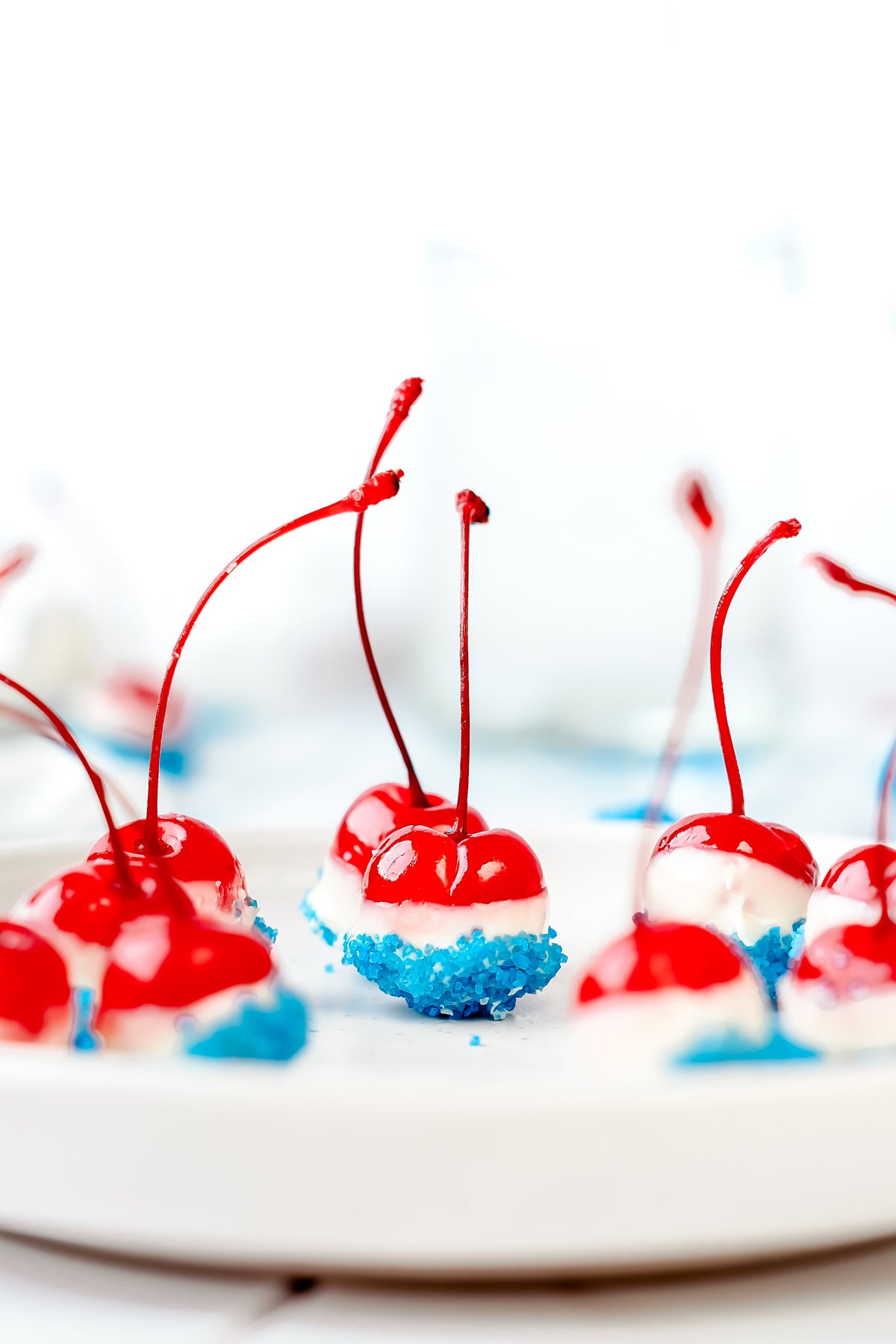 Red cherries dipped in white chocolate and blue sprinkles, arranged on a white plate for a festive treat.