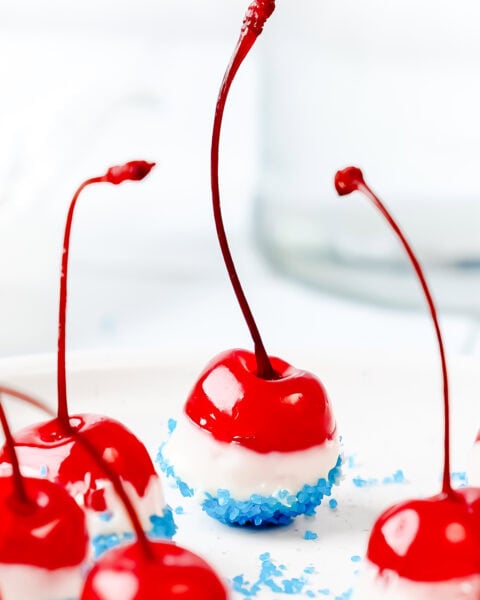 Bright red cherries dipped in white chocolate and blue sprinkles, arranged on a white surface.