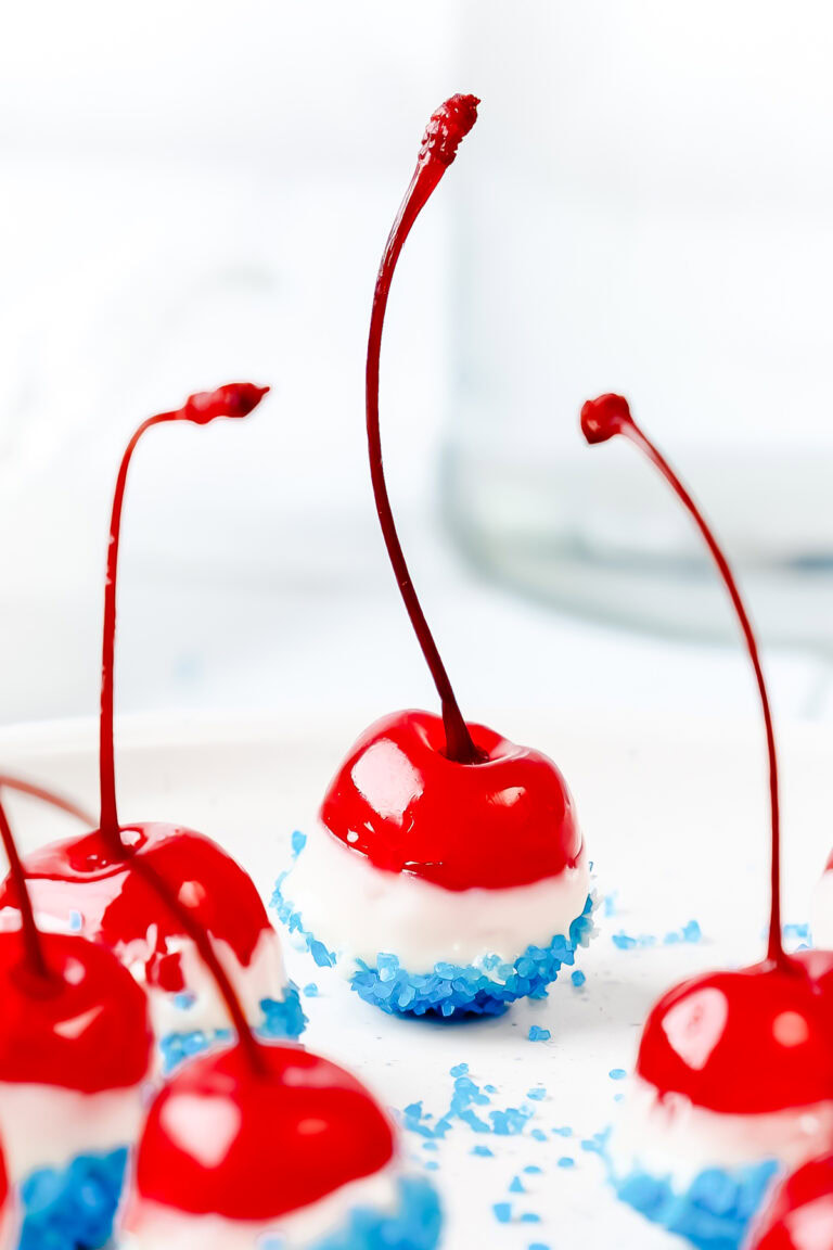 Bright red cherries dipped in white chocolate and blue sprinkles, arranged on a white surface.