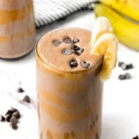 Chocolate banana smoothie in a glass, topped with banana slices and chocolate chips, with bananas in the background.